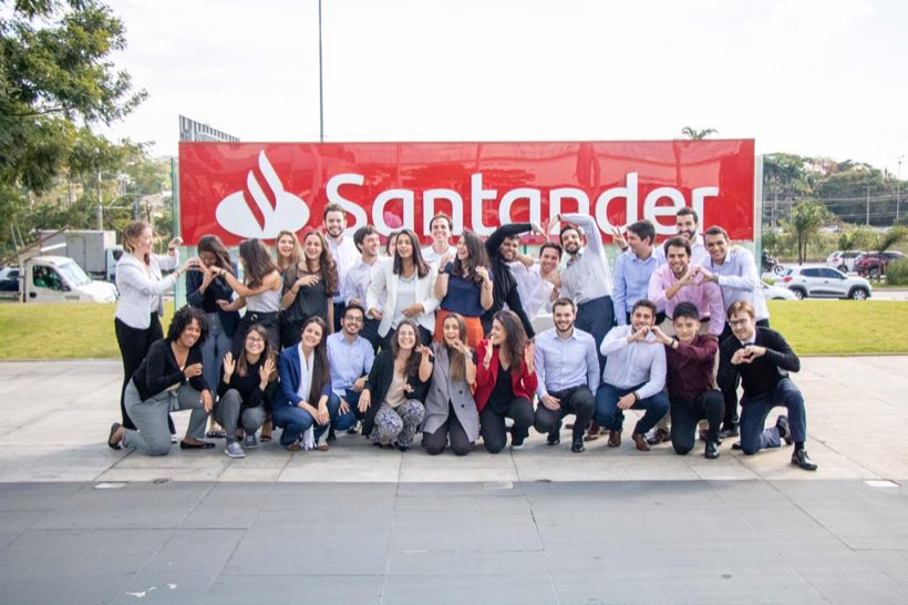 Turma de trainees do Santander Brasil reunida em frente ao letreiro da empresa.
